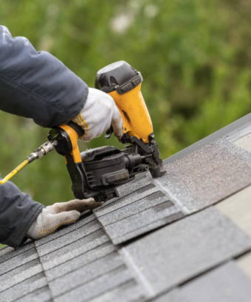 Unrecognizable roofer worker in uniform work wear using air or pneumatic nail gun and installing asphalt or bitumen tile on top of the roof under construction house